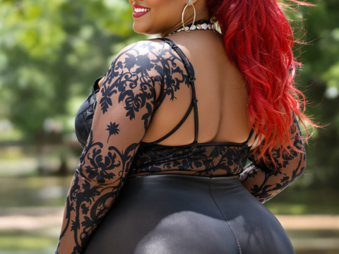 Portrait of a young woman with long red hair. she is standing in a park or garden with trees and greenery in the background. the woman is wearing a black lace bodysuit with a deep v-neckline and long sleeves. she has a pair of black high-waisted leggings that are tight-fitting and accentuate her curves. her hair is styled in loose waves and she is wearing large hoop earrings and a choker necklace. she appears to be smiling and looking over her shoulder at the camera. the overall mood of the image is confident and sultry.